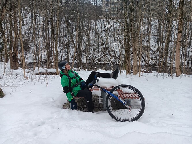 man lying on a bench in the snow with unicycle alonside