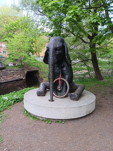 A metal sculpture of a elephant sitting down on its behind on a round concrete pedestal. The elephant has its front legs forward and the trunk reaching down into a hole in the pedestal. In the background is lush greenery of a park. It is also possible to make out part of the course of a waterway. There are some coloured buildings behind the trees. Resting againgst the chest of the elephant is a large red framed unicycle, with a tyre that has tan coloured sidewalls. The unicycle also has a disc brake.
